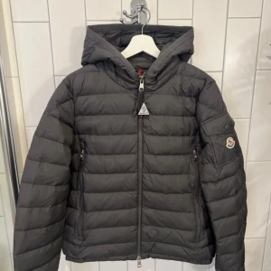 Grå Moncler dunjacka med huva ( Moncler Galion Down Jacket) - Snygg grå dunjacka från Moncler med huva och klassisk logga på ärmen. Jackan har quiltad design, dragkedja framtill och två fickor med dragkedja. Perfekt för kyliga dagar och ger en clean, modern look. Helt fräsch och ny med prislapp, finns en defekt på knappen där moncler loggan ligger så det går inte öppna eller stänga den osen så finns de en defekt på den triangel tagen som ligger fast på jackedkan, pris går att pruta och buda på.