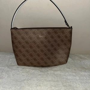 Brun axelväska från Guess - Snygg brun axelväska från Guess med klassiskt logomönster över hela väskan. Väskan har en enkel form, dragkedja upptill och en vit axelrem som ger en cool kontrast. Perfekt för dig som gillar stilrena accessoarer med lite lyxig känsla.