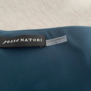 Mörkblå oneshoulder  jumpsuit Josie Natori - Supersnygg mörkblå klänning från Josie Natori med asymmetrisk oneshoulder-design och volangdetaljer. Klänningen har en figurnära passform upptill och utsvängd kjol, vilket ger en elegant siluett. Perfekt för dig som vill sticka ut med stil.