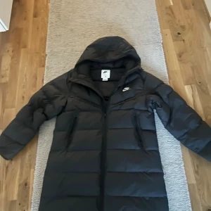 Svart lång pufferjacka från Nike - Säljer en svart lång pufferjacka från Nike med huva och Storm-FIT teknologi. Jackan har dragkedja framtill, två sidofickor med dragkedja och ett stilrent Nike-logga på bröstet. Perfekt för kalla dagar och har en clean, sportig vibe. Dessutom varm och bra inför vintern 