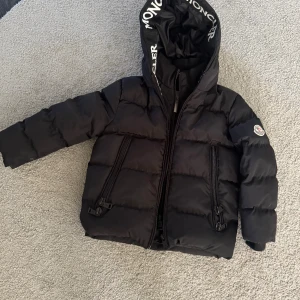 Svart Moncler pufferjacka med huva - Svart pufferjacka från Moncler med stor huva och tydlig Moncler-logga på huvkanten. Jackan har två snedställda fickor med dragkedja och ett broderat Moncler-märke på ärmen. Materialet är glansig polyester och jackan är quiltad för extra värme.