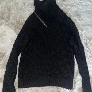 Svart polotröja från Armani Exchange - Snygg svart stickad polotröja från Armani Exchange med ribbad hög krage och diagonal dragkedja. Tröjan har ett diskret mönster, långa ärmar och en liten AX-logga vid nederkanten. Perfekt för dig som gillar stilrena och varma plagg.