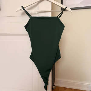 Mörkgrön body med smala axelband - Snygg mörkgrön body med snygg design och smala dubbla axelband. Modellen har en enkel, stilren form med hög skärning och raka linjer framtill. Perfekt för dig som gillar en clean look! 