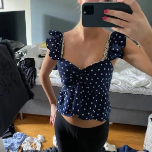 Mörkblå prickig blus med volang - Supersöt mörkblå blus med vita prickar och volangdetaljer på axlarna. Blusen har spetskant vid axelbanden, omlott framtill och en lätt croppad passform. Perfekt för dig som gillar romantisk och lekfull stil.