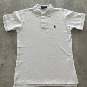 Vit pikétröja från Polo Ralph Lauren - Klassisk vit pikétröja från Polo Ralph Lauren i modellen Custom Slim Fit. Tröjan har korta ärmar, krage med knappar och den ikoniska broderade loggan på bröstet. Tillverkad i mjuk bomull som känns skön mot huden. Finns inga defekter, varken fläckar eller hål. Hör av er vid funderingar!