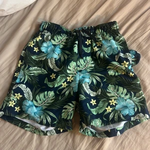 Blommiga badshorts från KappAhl - Snygga badshorts från KappAhl med tropiskt blommönster i blått, grönt och gult. Shortsen har elastisk midja med snörning och innerbyxa i mesh. Perfekta för stranden eller poolen med sin färgglada vibe och bekväma passform.