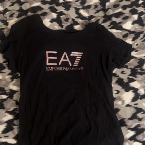 Svart EA7 Emporio Armani t-shirt - Svart t-shirt från EA7 Emporio Armani med metallicrosa logga framtill och tryck på ryggen. Klassisk rund halsringning och korta ärmar. Tillverkad i mjuk bomull för en skön känsla. Perfekt för dig som gillar stilrena och exklusiva plagg.