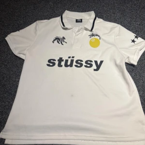 Vit Stüssy fotbollströja World Tour - Vit fotbollströja från Stüssy med svart tryck framtill och baktill, inklusive logga, lejon, stjärnor på ärmarna och World Tour-tryck med städer. Klassisk krage med svarta detaljer. Materialet är mjukt och andas, perfekt för streetwear.