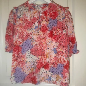 Blommig blus i rosa och lila toner - Färgstark blus med blommigt mönster i rosa, rött, lila och inslag av blått och gult. Modellen har trekvartsärmar med resår och knytning vid halsen. Luftigt material och avslappnad passform, perfekt för dig som gillar färg och mönster.