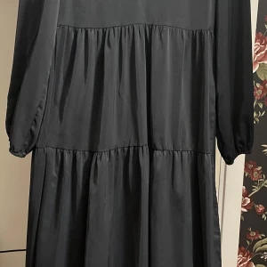 Svart Shein klänning - Svart lång klänning med volanger för en luftig passform. Klänningen har en lätt glansig yta och stängs med dragkedja i ryggen. Passar väldigt fint som en vardagsklänning eller kan kompletteras med en tjocktröja. En liten defekt orsakad av strykjärn på framsidan har gjort att det finns ett glansigt streck, men det syns knappt!