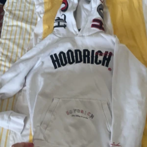 Vit hoodie från Hoodrich med tryck - Vit hoodie från Hoodrich med stor svart och röd logga på bröstet och detaljerat tryck på ryggen. Huva med tryck och känguruficka framtill. Mjuk och skön, perfekt för streetwear-stil.