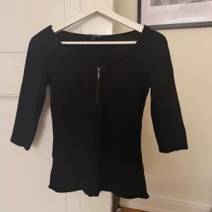 Svart topp med dragkedja  - Svart topp från Tally Weijl i storlek S med trekvartsärm och en snygg v-ringning framtill med dragkedja. Enkel och stilren design som passar till många olika outfits. Mjuk och skön bomullskänsla.