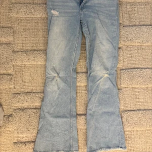 Bootcut ljusblå jeans från Zara - Säljer ett par ljusblå bootcut jeans från Zara med slitna detaljer vid knäna och rå kant längst ner. Klassisk femficksmodell med dragkedja och knapp framtill. Perfekt för dig som gillar en avslappnad och trendig look. Tveka inte på att höra av dig vid frågor eller intresse!