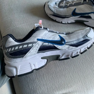 Nike sneakers vit och blå, strl 42.5 - Nike sneakers i vitt och marinblått med grå detaljer och en blå metallic swoosh. Skorna har snörning, robust sula och meshpartier för extra andningsförmåga. Perfekta för dig som gillar sportig stil och vill ha bekväma skor till vardagen.