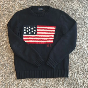 Mörkblå stickad tröja Polo Ralph Lauren - Mörkblå stickad tröja från Polo Ralph Lauren med amerikansk flagga framtill och RL-broderi. Klassisk rund halsringning och ribbade muddar vid ärmslut och nederkant. Perfekt för dig som gillar preppy stil och vill sticka ut lite extra.