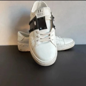 Valentino Rockstud vita sneakers - Snygga vita Valentino Rockstud sneakers i skinn med svart detalj och nitar över vristen. Klassisk rund tå, platt sula och snörning framtill. Ikoniska nitar längs sidorna ger en edgy vibe. Perfekta för dig som vill sticka ut med stil.