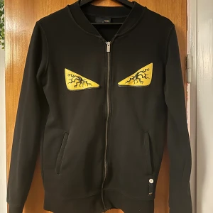 Svart hoodie från Fendi med ögon - Svart hoodie från Fendi med dragkedja och två fickor. På bröstet finns två gula applikationer som ser ut som ögon med svarta blixtliknande detaljer. Tröjan har lång ärm och en clean, modern look som sticker ut.