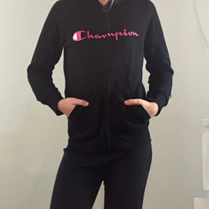 Svart hoodie från Champion med rosa tryck - Svart hoodie från Champion med dragkedja och huva. Framsidan har ett stort rosa Champion-tryck. Tröjan har långa ärmar, fickor på sidorna och är tillverkad i mjuk bomullsblandning. Perfekt för en sportig och avslappnad stil.
