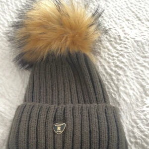 Rockandblue ribbstickad mössa med pälsboll - Mörkgrön ribbstickad mössa från Rockandblue med stor fluffig pälsboll i beige och svart upptill. Mössan har uppvikt kant och en liten metallbadge framtill med Rockandblue-logga. 