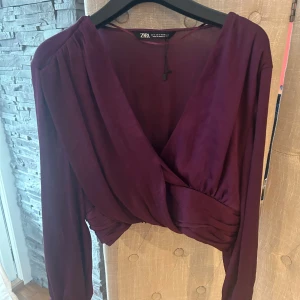 Vinröd omlottblus från Zara - Säljer en vinröd omlottblus från Zara i storlek S. Blusen har långa ärmar, djup v-ringning och ett snyggt omlottband i midjan. Materialet är lätt och har en silkeslen känsla, perfekt för att lyfta din outfit med en elegant touch.
