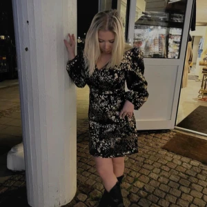 Svart glittrig kort klänning med v-ringning - Säljer en svart kort klänning med glittrigt mönster i guld. Klänningen har v-ringning, markerad midja och långa puffärmar. Perfekt för dig som vill sticka ut med en snygg och trendig look. Materialet känns festligt och skimrande.🥰