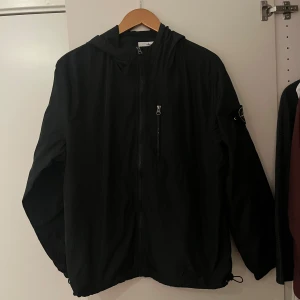 Svart vindjacka från Stone Island - Svart vindjacka från Stone Island med ikonisk patch på ärmen. Jackan har huva, dragkedja framtill, bröstficka med dragkedja och justerbar nederkant. Tillverkad i lätt polyester som passar perfekt för blåsiga dagar. Snygg och stilren design med normal passform.