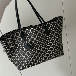 Malene Birger Svart shoppingväska med rutigt mönster - Stor och rymlig shoppingväska från Marlene Birger i svart med beige rutigt mönster. Väskan har två långa svarta handtag, guldfärgad stängning och en snygg tag-detalj. Materialet är slitstark syntet och designen är både modern och klassisk. Ordinarie 2799kr, Pris 2000kr