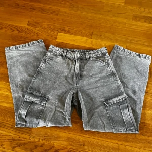 Grå cargopants i jeansmaterial - Säljer ett par grå cargopants i tvättad jeanslook med breda ben och stora fickor på sidorna. Byxorna har hög midja, raka ben och cool streetstil. Perfekta för dig som gillar avslappnad vibe och praktiska detaljer.