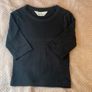 Svart tajt basic topp från H&M - En svart, enkel topp från H&M med rund halsringning och trekvartsärm. Storlek 134-140 men är jätte stretchig. Modellen är slim och sitter snyggt mot kroppen. Tillverkad i mjuk bomull som är skön att bära. Perfekt till jeans eller kjol för en clean look.