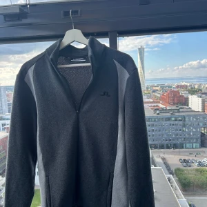 Mörkgrå ziptröja från J.Lindeberg - Snygg mörkgrå ziptröja från J.Lindeberg med svart logga på bröstet och nacken. Tröjan har hel dragkedja, hög krage och är tillverkad i ett mjukt material. Perfekt för lager-på-lager och stilren look.