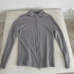 Grå kofta med dragkedja från j.lindeberg - Stilren grå kofta från j.lindeberg med hel dragkedja framtill och klassisk krage. Långärmad och tillverkad i mjukt material som passar perfekt till lager-på-lager. Enkel och clean look som funkar till det mesta.