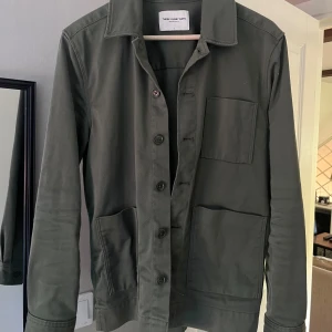 Grön overshirt från These Glory Days - Stilren grön overshirt från These Glory Days med klassisk krage, tre stora fickor framtill och knäppning med knappar. Jackan är i bomull och har en rak, normal passform med långa ärmar. Perfekt för lager-på-lager och enkel att matcha.