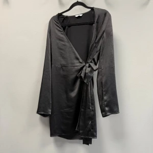 Svart omlottklänning i satin - Snygg svart kort omlottklänning från Envii i glansigt satinmaterial. Klänningen har långa ärmar och knytdetalj i midjan som ger en schysst siluett, passar för både S och M💗💗