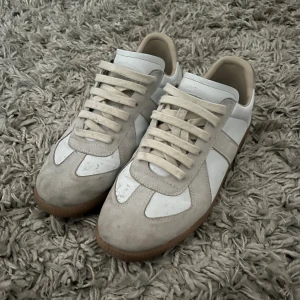 Maison margiela skor  - Säljer ett par stilrena sneakers i vitt och beige med snygga mockadetaljer och klassisk gummisula. Skorna har snörning och en låg profil som passar till många olika outfits. Perfekta för dig som gillar minimalistisk och clean stil.