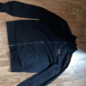 Svart EA7 Emporio Armani tracksuit - Svart tracksuit från EA7 Emporio Armani med guldfärgad logga på både jacka och byxa. Setet har dragkedja på jackan, muddar vid ärmslut och benslut samt sidofickor. Tillverkad i mjuk polyester för en sportig och clean look. Koftan använt än del men är i bra skick är lite skada vid höger nedre sida men inget illa, byxorna knappt använda använt kanske 5 ganger max. Jag säljer dessa då det inte passar mig längre. Om man bara vill köpa till exempel bara koftan eller bara byxorna så skriv till mig(: