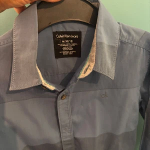 Blå skjorta från Calvin Klein Jeans - Snygg blå skjorta från Calvin Klein Jeans med diskret rutigt mönster i olika blå nyanser. Skjortan har klassisk krage, knäppning framtill och broderad ck-logga på bröstet. Tillverkad i bomull och polyester, perfekt för en stilren look.