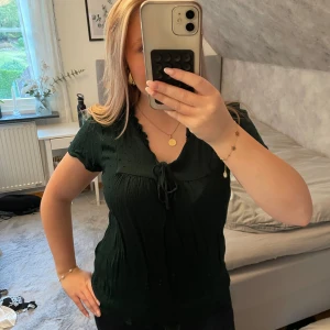 Mörkgrön blus med knyt från JDY - Supersöt mörkgrön blus från JDY med små prickar i tyget, v-ringning och knyt framtill. Blusen har korta ärmar och volangkant vid halsen. Perfekt att styla med jeans eller kjol för en chill look.