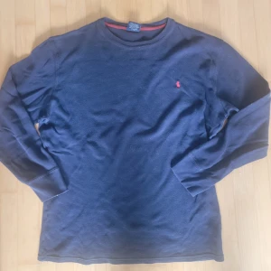 Marinblå långärmad tröja Polo Ralph Lauren - Marinblå långärmad tröja från Polo Ralph Lauren i 100% bomull. Klassisk rund hals och diskret röd broderad logga på bröstet. Tröjan har ribbade muddar och en enkel, stilren design som funkar till många olika looks.