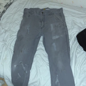 Grå Carhartt cargopants relaxed fit - Grå cargopants från Carhartt i relaxed fit med slitna detaljer och lagningar. Byxorna har stora bakfickor, extra benficka och klassisk Carhartt-logga på bakfickan. Perfekta för en avslappnad och streetig stil. Har dock vit färg pläckar, som kanske är möjligt att ta bort.