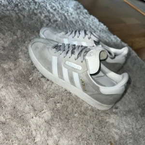 Adidas Gazelle grå/vita sneakers - Säljer ett par Adidas Gazelle sneakers i grå mocka med vita detaljer och klassiska tre ränder på sidorna. Skorna har snörning, platt sula och rund tå. Guldtexten 'GAZELLE' på sidan ger en snygg touch. Perfekta för dig som gillar stilrena och tidlösa sneakers.