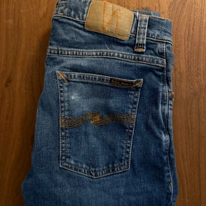 Nudie Jeans W29 L30 - Säljer ett par klassiska blå jeans från Nudie Jeans med snygga slitningar och kontrastsömmar. Modellen har fem fickor, orange broderi på bakfickorna och slim passform