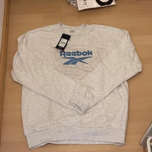 Grå Reebok sweatshirt med blå logga - Snygg ljusgrå sweatshirt från Reebok med broderad blå logga framtill. Tröjan har rund hals, långa ärmar och ribbade muddar vid ärmslut och nederkant. Tillverkad i mjuk bomullsmix, perfekt för chill eller träning. Tröjan är helt ny direkt från asos. 