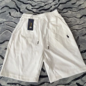 Vita shorts från Polo Ralph Lauren - Snygga vita shorts från Polo Ralph Lauren med klassisk broderad logga i svart på ena benet. Modellen har elastisk midja med snörning och två sidofickor. Perfekta för sommaren och riktigt clean look. 