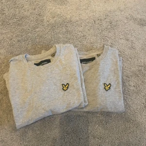 Grå t-shirt från Lyle & Scott - Säljer två stilrena grå t-shirts från Lyle & Scott i 100% ekologisk bomull. Klassisk passform med korta ärmar och rund hals. Bröstet pryds av den ikoniska gula örn-loggan. Perfekt basic-plagg med mjuk känsla och enkel design.