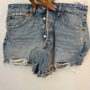 Ljusblåa jeansshorts - Säljer ett par högmidjade ljusblå jeansshorts med slitna detaljer och fransig kant. Klassisk femficksmodell med knappar framtill och bälteshällor. Perfekta för varma dagar och en avslappnad look.