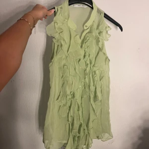 Ljusgrön volangblus från Mango - Supersöt ljusgrön blus från MNG med volanger framtill och knappar hela vägen. Ärmlös modell i ett tunt, skirt tyg som ger en luftig känsla. Perfekt för dig som gillar romantiska detaljer och vill sticka ut med en unik topp.