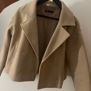 Beige kort kavajjacka från Bik Bok - Kort, beige kavajjacka med dubbelknäppning och breda slag. Jackan har två fickor framtill, dekorativa knappar bak och är helfodrad. Perfekt för dig som gillar stilren och trendig look.