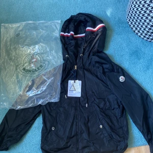 Svart vindjacka från Moncler med huva - Svart vindjacka från Moncler med huva och dragsko, dekorerad med röd och vit rand på huvan. Jackan har dragkedja framtill, två fickor med tryckknappar och Moncler-logga på ärmen. Perfekt för blåsiga dagar och snygg streetstil.