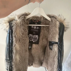 Beige pälsväst från Navahoo - Snygg beige pälsväst från Navahoo med fluffig fuskpäls och svarta glansiga paneler i syntetmaterial. Västen har huva med pälsdetaljer och snörning i midjan för en justerbar passform. Perfekt för att ge din outfit en extra edge.