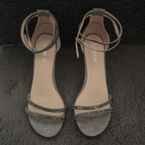 Grå sandaler med klack från Catwalk - Snygga grå sandaler från Catwalk med smala remmar dekorerade med glittriga detaljer och en elegant ankelrem med spänne. Skorna har öppen tå och en smal klack. Endast använda 1 gång. Kan både skickas eller hämtas på plats.